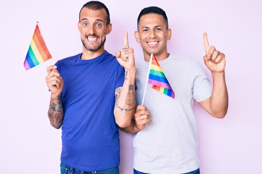 Young gay couple of two men holding rainbow lgbtq flags together smiling with an idea or question pointing finger with happy face, number one