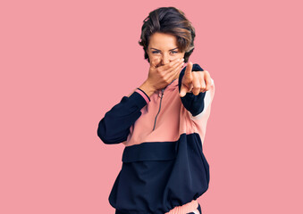 Young beautiful woman wearing sportswear laughing at you, pointing finger to the camera with hand over mouth, shame expression