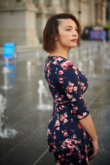 Beautiful biracial woman in floral print dress poses among fountains in city plaza