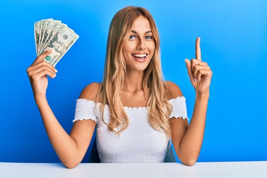 Beautiful blonde young woman holding 20 dollars banknotes smiling with an idea or question pointing finger with happy face, number one