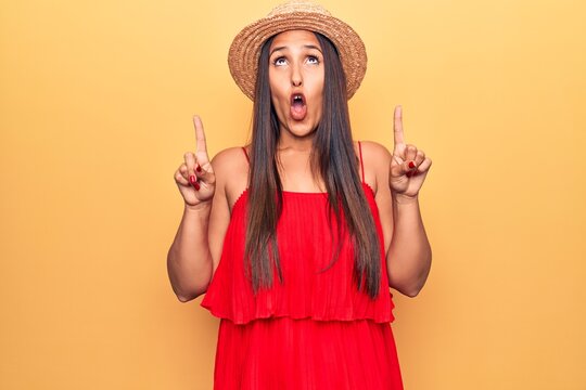 Young beautiful brunette woman wearing summer hat and dress amazed and surprised looking up and pointing with fingers and raised arms.