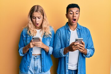 Young interracial couple using smartphone afraid and shocked with surprise and amazed expression, fear and excited face.