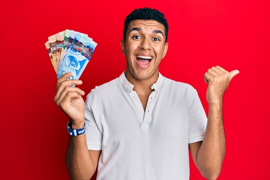 Young arab man holding canadian dollars pointing thumb up to the side smiling happy with open mouth