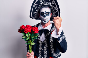 Young man wearing mexican day of the dead costume holding flowers annoyed and frustrated shouting with anger, yelling crazy with anger and hand raised