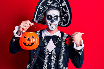 Young man wearing mexican day of the dead costume holding pumpkin pointing thumb up to the side smiling happy with open mouth