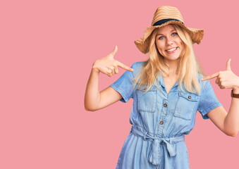 Fototapeta premium Young beautiful blonde woman wearing summer hat and dress looking confident with smile on face, pointing oneself with fingers proud and happy.