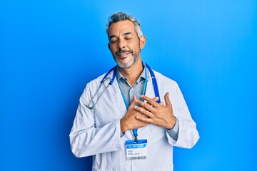Middle age grey-haired man wearing doctor uniform and stethoscope smiling with hands on chest with...