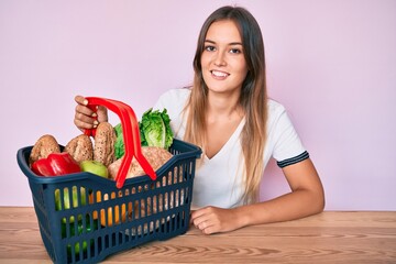 Fototapeta na wymiar Beautiful caucasian woman holding supermarket shopping basket looking positive and happy standing and smiling with a confident smile showing teeth