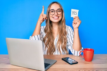 Young beautiful blonde woman holding yes reminder sitting on the table smiling with an idea or...