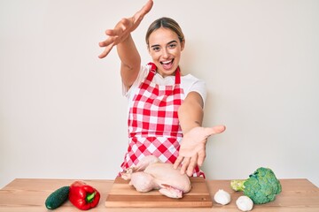 Fototapeta premium Young beautiful blonde woman wearing apron cooking chicken looking at the camera smiling with open arms for hug. cheerful expression embracing happiness.