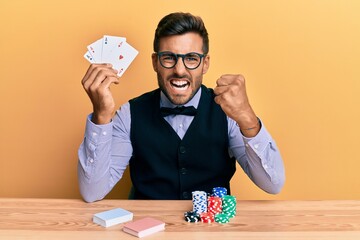 Handsome hispanic croupier man sitting on the table with poker chips and cards annoyed and frustrated shouting with anger, yelling crazy with anger and hand raised