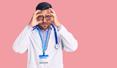 Young hispanic man wearing doctor uniform and stethoscope trying to open eyes with fingers, sleepy and tired for morning fatigue