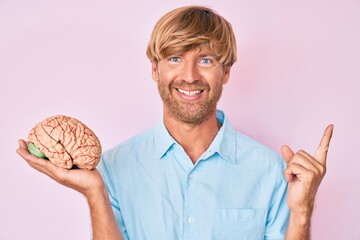 Young blond man holding brain smiling happy pointing with hand and finger to the side