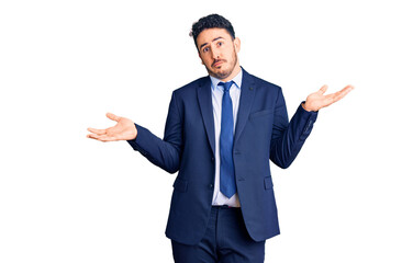 Young hispanic man wearing business clothes clueless and confused expression with arms and hands raised. doubt concept.