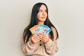 Young beautiful hispanic girl holding swiss franc banknotes smiling looking to the side and staring away thinking.