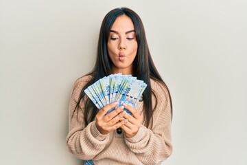 Young beautiful hispanic girl holding south african 100 rand banknotes making fish face with mouth and squinting eyes, crazy and comical.