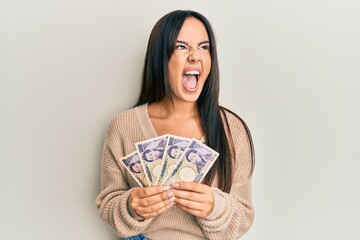 Young beautiful hispanic girl holding japanese yen banknotes angry and mad screaming frustrated and furious, shouting with anger. rage and aggressive concept.