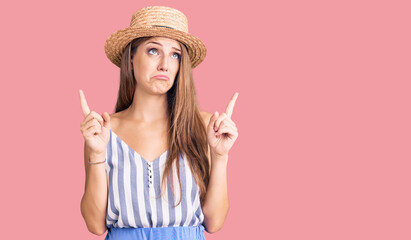 Young beautiful blonde woman wearing summer hat pointing up looking sad and upset, indicating direction with fingers, unhappy and depressed.