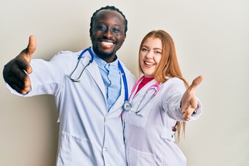 Young interracial couple wearing doctor uniform and stethoscope looking at the camera smiling with...