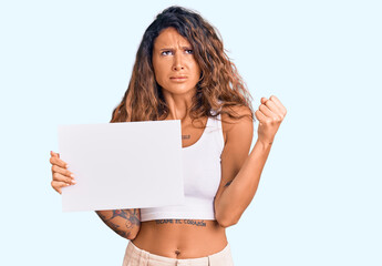 Young hispanic woman with tattoo holding blank empty paper annoyed and frustrated shouting with anger, yelling crazy with anger and hand raised