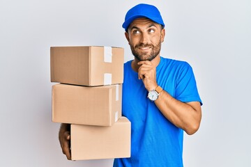 Handsome man with beard wearing courier uniform holding delivery packages with hand on chin thinking about question, pensive expression. smiling and thoughtful face. doubt concept.