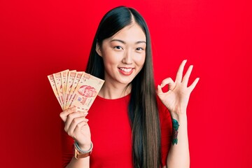 Young chinese woman holding mexican pesos doing ok sign with fingers, smiling friendly gesturing excellent symbol