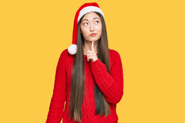 Young chinese woman wearing christmas hat thinking concentrated about doubt with finger on chin and looking up wondering