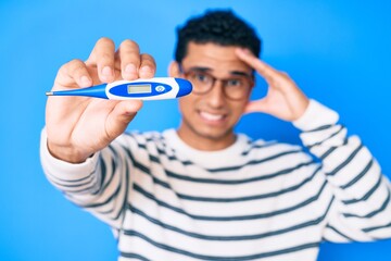 Naklejka premium Young handsome hispanic man holding thermometer stressed and frustrated with hand on head, surprised and angry face