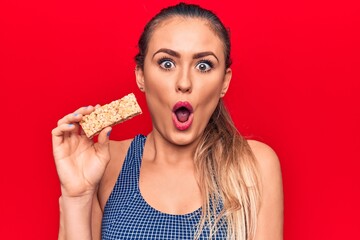Young beautiful blonde woman eating muesli cereal bar over isolated red background scared and...