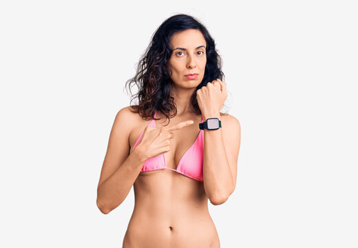 Young beautiful hispanic woman wearing bikini in hurry pointing to watch time, impatience, looking at the camera with relaxed expression