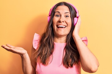 Young beautiful brunette woman listening to music using headphones over yellow background celebrating achievement with happy smile and winner expression with raised hand