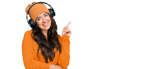 Beautiful brunette young woman listening to music using headphones with a big smile on face, pointing with hand and finger to the side looking at the camera.