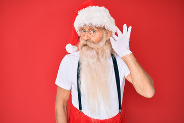 Old senior man with grey hair and long beard wearing white t-shirt and santa claus costume smiling with hand over ear listening an hearing to rumor or gossip. deafness concept.