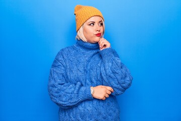 Young beautiful blonde plus size woman wearing blue casual turtleneck sweater and wool hat thinking...