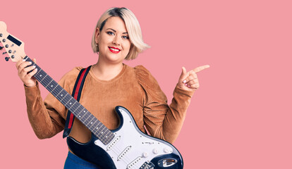 Fototapeta premium Young blonde plus size woman playing electric guitar smiling happy pointing with hand and finger to the side