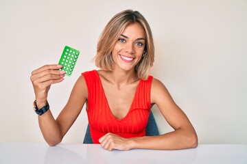 Beautiful caucasian woman holding birth control pills looking positive and happy standing and...