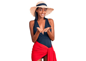Young african american woman wearing swimsuit and summer hat smiling with hands palms together receiving or giving gesture. hold and protection