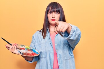 Young plus size woman holding paintbrush and palette pointing with finger to the camera and to you,...
