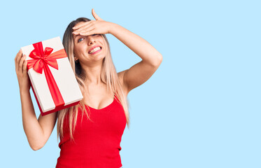 Young beautiful blonde woman holding gift stressed and frustrated with hand on head, surprised and angry face