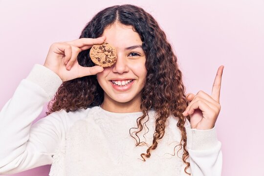 Beautiful kid girl with curly hair holding cookie smiling happy pointing with hand and finger to the side