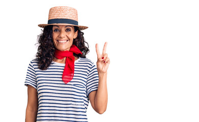 Middle age beautiful woman wearing casual striped t shirt and summer hat smiling with happy face winking at the camera doing victory sign. number two.