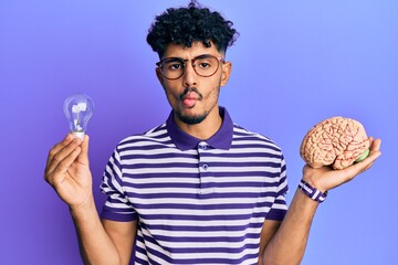 Young arab handsome man holding brain and lightbulb for inspiration and idea making fish face with...