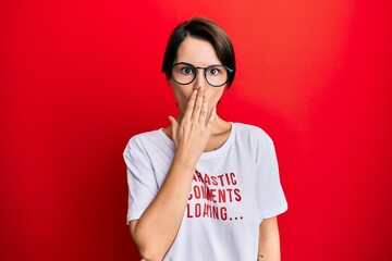 Young brunette woman with short hair wearing sarcastic comments loading t-shirt covering mouth with hand, shocked and afraid for mistake. surprised expression