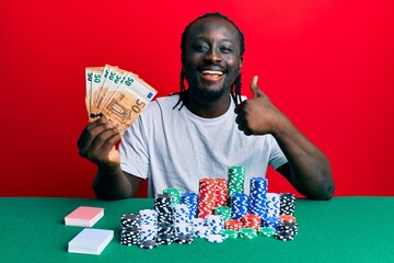 Handsome young black man playing poker holding 50 euros banknotes smiling happy and positive, thumb up doing excellent and approval sign