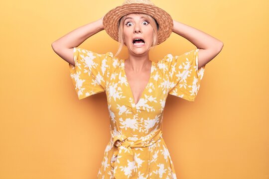 Beautiful blonde woman on vacation wearing summer hat and dress over yellow background Crazy and scared with hands on head, afraid and surprised of shock with open mouth