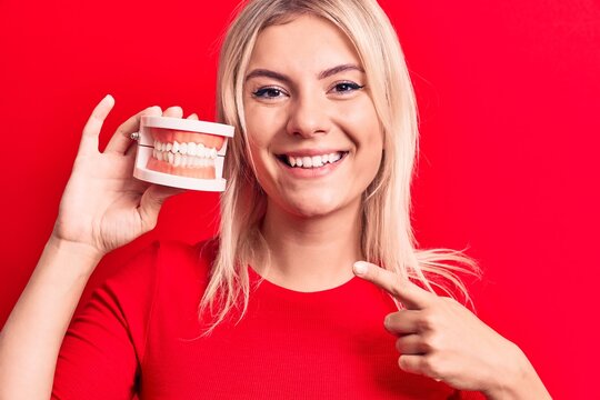 Young Beautiful Blonde Woman Holding Denture Teeth Standing Over Isolated Red Background Smiling Happy Pointing With Hand And Finger