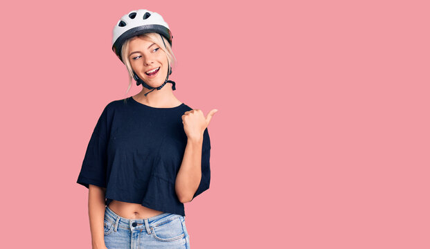 Young beautiful blonde woman wearing bike helmet smiling with happy face looking and pointing to the side with thumb up.