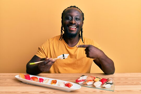 Handsome young black man eating sushi sitting on the table smiling happy pointing with hand and finger