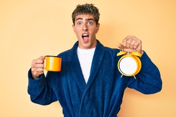 Fototapeta premium Young handsome man wearing bathrobe holding cup of coffee and alarm clock celebrating crazy and amazed for success with open eyes screaming excited.