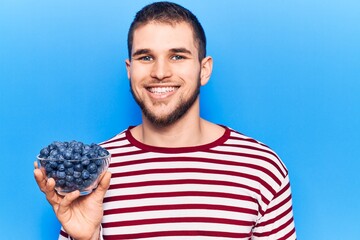 Young handsome man holding bowl with blueberries looking positive and happy standing and smiling with a confident smile showing teeth